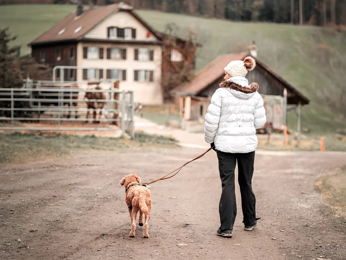 Eine Frau in weisser Jacke führt einen kleinen Hund an der Leine auf einem Feldweg zu einem Bauernhof
