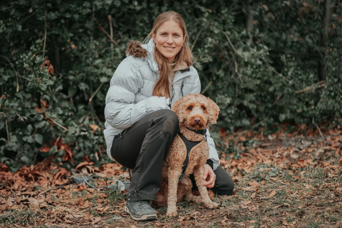 Olivia Döring hockt im Herbstwald neben ihrem Pudelmix und lächelt in die Kamera